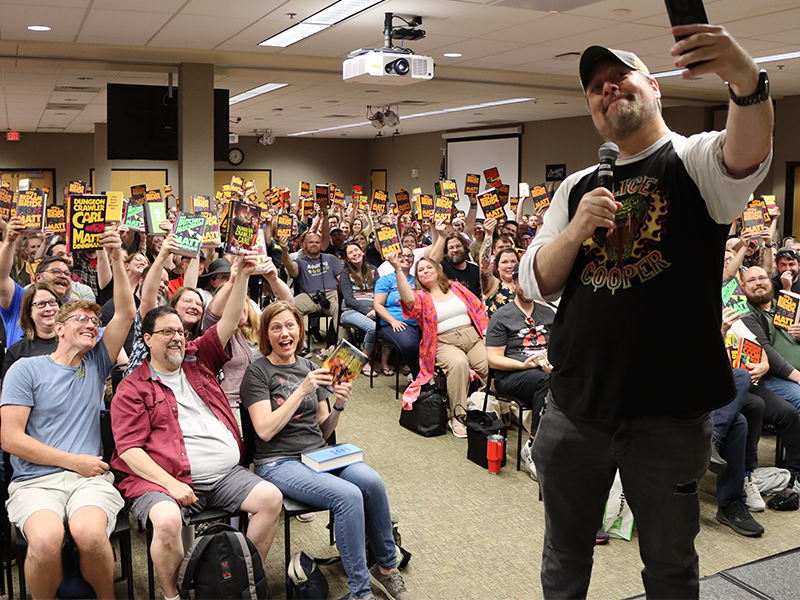 a library author event with a room full of people holding the authors book up for a photo