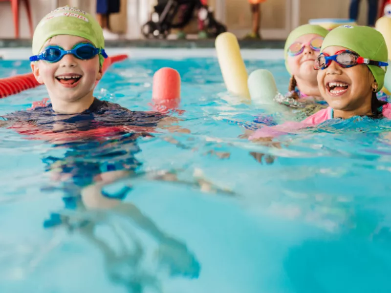 children swimming in pool with swim caps, goggles, and pool noodles